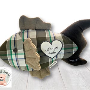 May include: A plush fish toy made from a green, white, and blue plaid fabric with solid brown fins and tail. A white heart-shaped patch on the side features the embroidered text "love you Grandpa". A small black button serves as the fish eye.