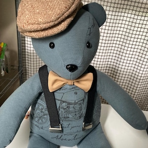 May include: A blue fabric teddy bear wearing a tan tweed flat cap, a tan bow tie, and black suspenders with silver hardware. The bear's torso features a graphic print of a motorcycle and the text "Start your own Adventure" in black ink.