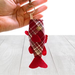 May include: A fish-shaped keychain featuring a red and white plaid fabric body with a single brown button eye. The fish has solid red felt fins and a red felt tail, attached to a silver-toned metal split ring by a short, thick white cord.