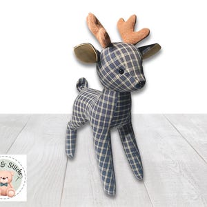 May include: A stuffed deer toy crafted from navy blue and white plaid fabric. The deer features light brown textured antlers, dark brown ears, and small black bead eyes. A circular logo with a teddy bear and the text Hugs & Stitches is visible.