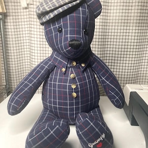 May include: A stuffed teddy bear wearing a plaid cap and shirt. The bear is navy blue with a red and white plaid pattern. The bear has a black nose and button eyes. The word "Grampy" is embroidered on one foot with a red heart.