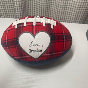 May include: A plush football made of red and black plaid fabric with white ribbon laces. A white heart-shaped patch is centered on the front, featuring the black embroidered text "Love, Grandpa" in a cursive and print font combination.