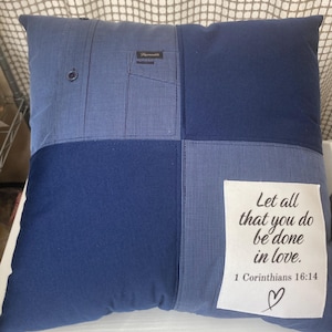 May include: A square throw pillow constructed from blue fabric patches, including a section of a button-down shirt with a pocket. A white rectangular patch is sewn on the front with the black text: Let all that you do be done in love. 1 Corinthians 16:14, and a small heart icon.