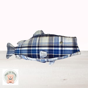 May include: A fish-shaped pillow made of blue, white, and navy plaid fabric. The pillow has fins, a tail, and a button eye. The pillow is on a white wooden surface. The logo "Hugs & Stitches" is in the bottom left corner.