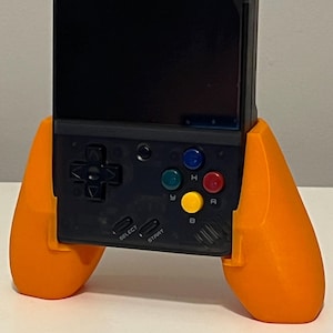 Peut inclure: Une console de jeu vidéo portable noire avec des manettes orange attachées. La console possède un écran, un pavé directionnel, quatre boutons et les mots "Select" et "Start" imprimés dessus.