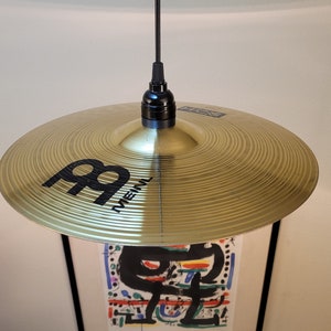 Op de afbeelding: Een gouden bekkens met een zwart "Paiste" logo en een zwart snoer dat hangt aan een zwarte lampfitting.