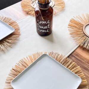 May include: A brown glass vase with the text "home sweet home" written on it, filled with dried flowers. The vase is sitting on a woven straw placemat. There are three other woven straw placemats with white square plates on top of them.