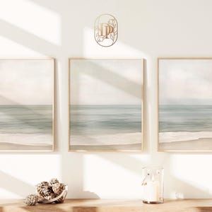 Peut inclure: Trois impressions encadrées d'un paysage de plage avec une vue sereine sur l'océan dans des tons de bleu, beige et blanc. Un petit bougeoir en verre avec une bougie allumée et un bol de pommes de pin sont sur une surface en bois.