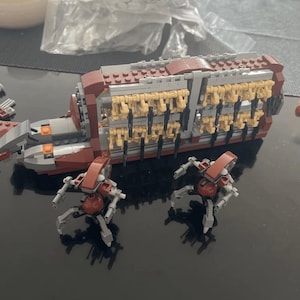 Puede incluir: Un modelo de Lego de un vehículo de transporte rojo y marrón, lleno de figuras marrones. Dos pequeñas figuras de robot rojas y grises están frente al vehículo, sobre una superficie oscura.
