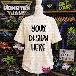 May include: A cream-colored t-shirt with the text "YOUR DESIGN HERE" in black. The shirt is displayed on a mannequin. The background features Monster Jam logos, toy trucks, and the text "DRIVE IT CRUSH IT LOVE IT".