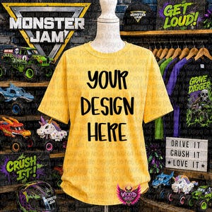 May include: A yellow t-shirt with the text "YOUR DESIGN HERE" in black. The shirt is displayed on a mannequin. The background includes Monster Jam graphics and toy trucks.