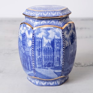 May include: A blue and white ceramic jar with a lid, featuring a detailed illustration of Ripon Cathedral. The jar has gold trim and a hexagonal shape. The design is reminiscent of classic blue and white pottery.