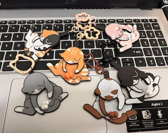 May include: A collection of cat-shaped keychains in various colors, including orange, gray, black, and pink. Each keychain features a sleeping cat design with a small ball detail. Some have flower-shaped attachments.