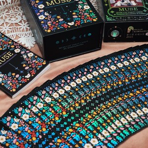 K&ouml;nnte beinhalten: Ein Set "Muse Cat Tarot Cards" mit floralen und himmlischen Designs. Die schwarzen Schachteln und Kartenr&uuml;ckseiten zeigen bunte Blumen und das Wort "MUSE". Die Karten sind aufgef&auml;chert und zeigen komplizierte Muster. Der Text "MUSE CAT TAROT CARDS BY KIMDAJEONG" ist sichtbar.