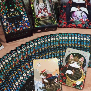 K&ouml;nnte beinhalten: Ein Tarotkartenspiel mit Katzenthema, mit dem Wort "MUSE" auf der Schachtel und den Karten. Das Deck enth&auml;lt farbenfrohe Blumendesigns, Katzenillustrationen und den Text "THE FOOL" auf einer Karte. Die Schachtel ist schwarz mit farbenfrohen Blumendesigns.