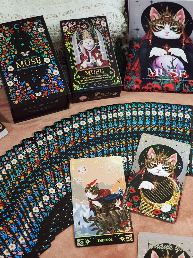 K&ouml;nnte beinhalten: Ein Tarotkartenspiel mit Katzenthema, mit dem Wort "MUSE" auf der Schachtel und den Karten. Das Deck enth&auml;lt farbenfrohe Blumendesigns, Katzenillustrationen und den Text "THE FOOL" auf einer Karte. Die Schachtel ist schwarz mit farbenfrohen Blumendesigns.