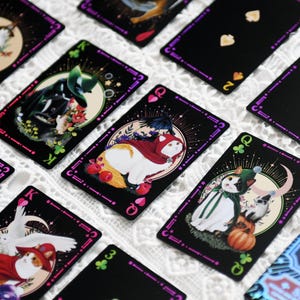 Könnte beinhalten: Schwarze Spielkarten mit lila Rändern und Illustrationen von Katzen in verschiedenen Kostümen. Die Karten zeigen Farben und Bildkarten mit detaillierten Kunstwerken und einem skurrilen Thema. Die Karten sind auf einer weißen, strukturierten Oberfläche angeordnet.