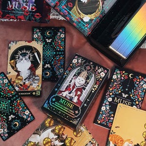 K&ouml;nnte beinhalten: Eine Sammlung von "MUSE" Katzen-Tarotkarten mit verschiedenen Illustrationen. Die Karten zeigen Katzen in k&ouml;niglicher Kleidung, umgeben von floralen und himmlischen Designs. Die Box und die Karten haben einen schwarzen Hintergrund mit goldenen Akzenten.