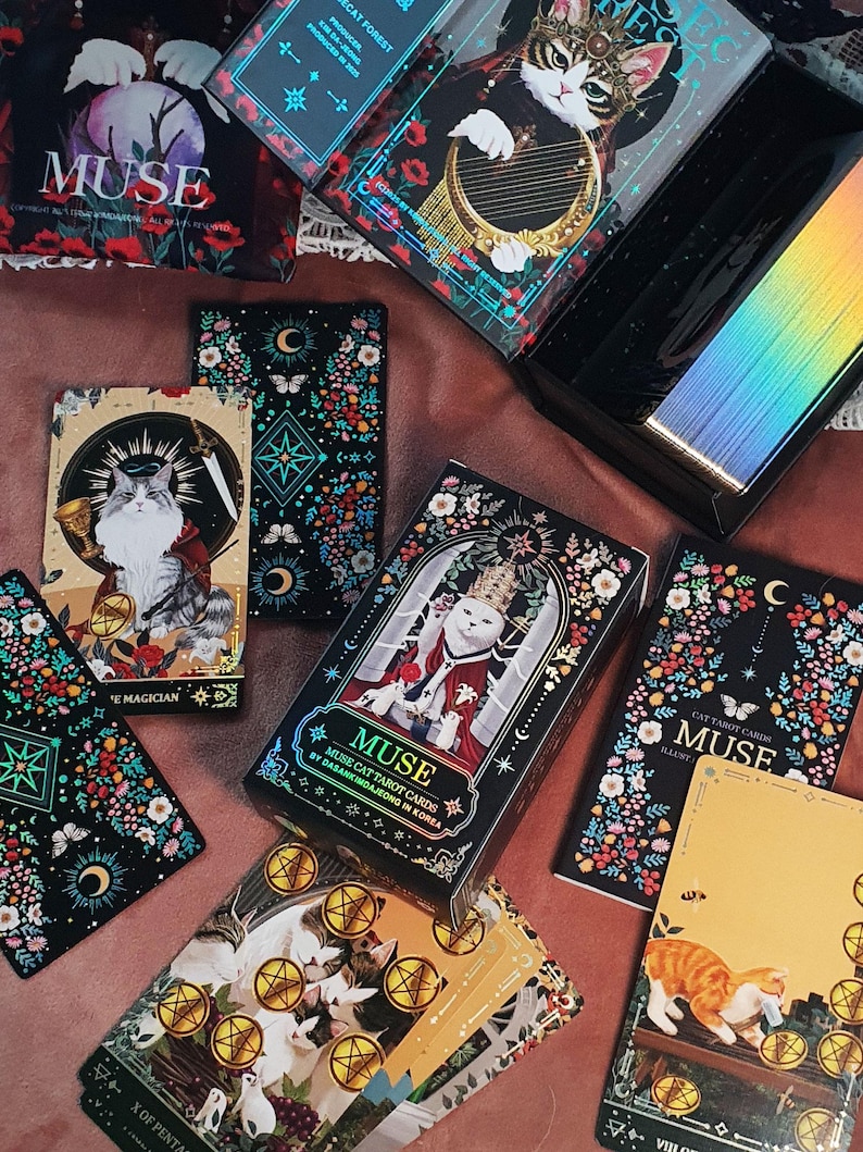 K&ouml;nnte beinhalten: Eine Sammlung von "MUSE" Katzen-Tarotkarten mit verschiedenen Illustrationen. Die Karten zeigen Katzen in k&ouml;niglicher Kleidung, umgeben von floralen und himmlischen Designs. Die Box und die Karten haben einen schwarzen Hintergrund mit goldenen Akzenten.