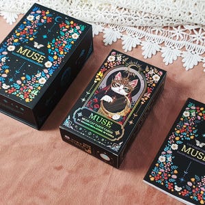 K&ouml;nnte beinhalten: Drei schwarze Tarotkartenboxen mit Blumen- und Schmetterlingsdesigns. Auf den Boxen steht "MUSE" und "CAT TAROT CARDS". Eine Box zeigt eine Katze, die eine Harfe spielt. Die Boxen befinden sich auf einer rosa Oberfl&auml;che.