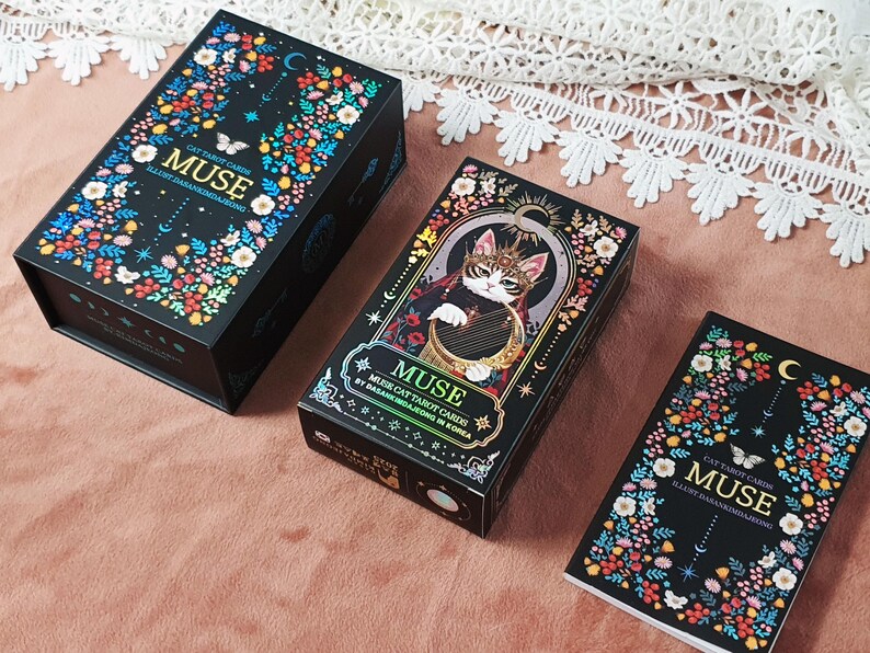 K&ouml;nnte beinhalten: Drei schwarze Tarotkartenboxen mit Blumen- und Schmetterlingsdesigns. Auf den Boxen steht "MUSE" und "CAT TAROT CARDS". Eine Box zeigt eine Katze, die eine Harfe spielt. Die Boxen befinden sich auf einer rosa Oberfl&auml;che.