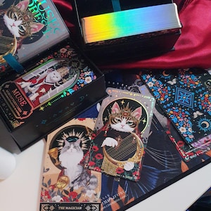 K&ouml;nnte beinhalten: Ein Set aus Tarotkarten und -boxen mit Katzenillustrationen. Die Karten und Boxen haben einen schwarzen Hintergrund mit farbenfrohen Blumen- und Himmelsmustern. Die Karten zeigen Katzen in k&ouml;niglicher Kleidung, mit dem Text "MUSE" und "MUSIC FOREST".