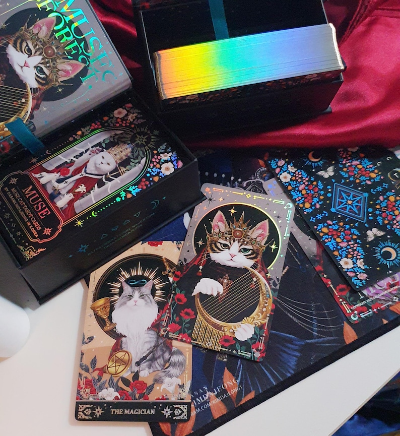 K&ouml;nnte beinhalten: Ein Set aus Tarotkarten und -boxen mit Katzenillustrationen. Die Karten und Boxen haben einen schwarzen Hintergrund mit farbenfrohen Blumen- und Himmelsmustern. Die Karten zeigen Katzen in k&ouml;niglicher Kleidung, mit dem Text "MUSE" und "MUSIC FOREST".