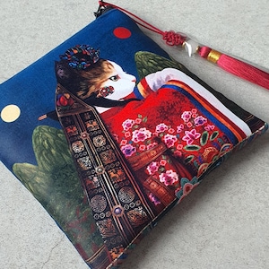 Könnte beinhalten: Quadratische, blaue Tasche mit einer Katze in traditioneller asiatischer Kleidung, vor einem Nachthimmel mit Mond und rotem Punkt. Die Katze trägt einen floralen Kopfschmuck und ein rotes Gewand mit floralen Mustern. Eine rote Quaste hängt oben.
