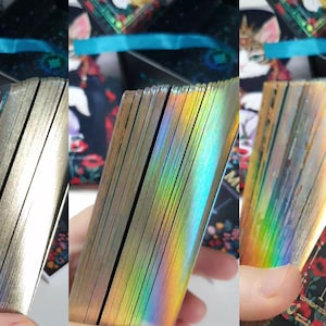 K&ouml;nnte beinhalten: Ein Tarotkartenspiel mit goldfarbenen Kanten, gehalten in einer Hand. Die Kanten der Karten reflektieren einen Regenbogen an Farben. Die Karten zeigen eine Katzenillustration mit floralen und himmlischen Elementen.