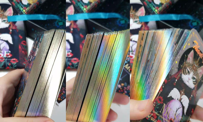 K&ouml;nnte beinhalten: Ein Tarotkartenspiel mit goldfarbenen Kanten, gehalten in einer Hand. Die Kanten der Karten reflektieren einen Regenbogen an Farben. Die Karten zeigen eine Katzenillustration mit floralen und himmlischen Elementen.