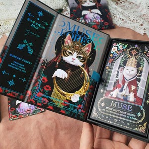 K&ouml;nnte beinhalten: Ein Tarotkartenset mit einer Katze mit Krone und Harfe, betitelt "Musecat Forest 2025". Die Box und das Buch haben Blumen- und Schl&uuml;sselmuster, mit dem Wort "MUSE". Das Set enth&auml;lt eine Box und ein Buch.