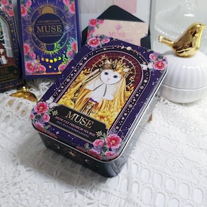 Könnte beinhalten: Rechteckige Blechdose mit Katzenillustration, dem Wort "MUSE" und floralen Akzenten. Die Dose hat einen dunkelvioletten Hintergrund und abgerundete Kanten. Weitere Dosen und eine goldene Vogelfigur sind ebenfalls sichtbar.