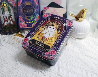 Könnte beinhalten: Rechteckige Blechdose mit Katzenillustration, dem Wort "MUSE" und floralen Akzenten. Die Dose hat einen dunkelvioletten Hintergrund und abgerundete Kanten. Weitere Dosen und eine goldene Vogelfigur sind ebenfalls sichtbar.