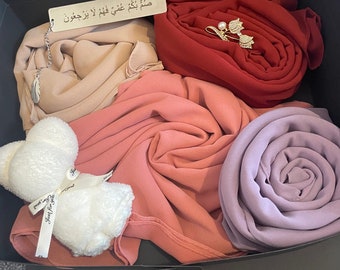 May include: A gift box filled with five scarves in shades of pink, purple, and red. The box also includes a small white plush toy with a tag that reads "Just for You". A small silver tag with Arabic text is also included.