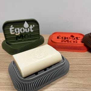 Peut inclure: Un porte-savon gris avec un savon, avec un design nervuré. Le porte-savon porte le texte "Égout' Moi" en blanc. D'autres porte-savons verts, oranges et marrons sont visibles en arrière-plan.