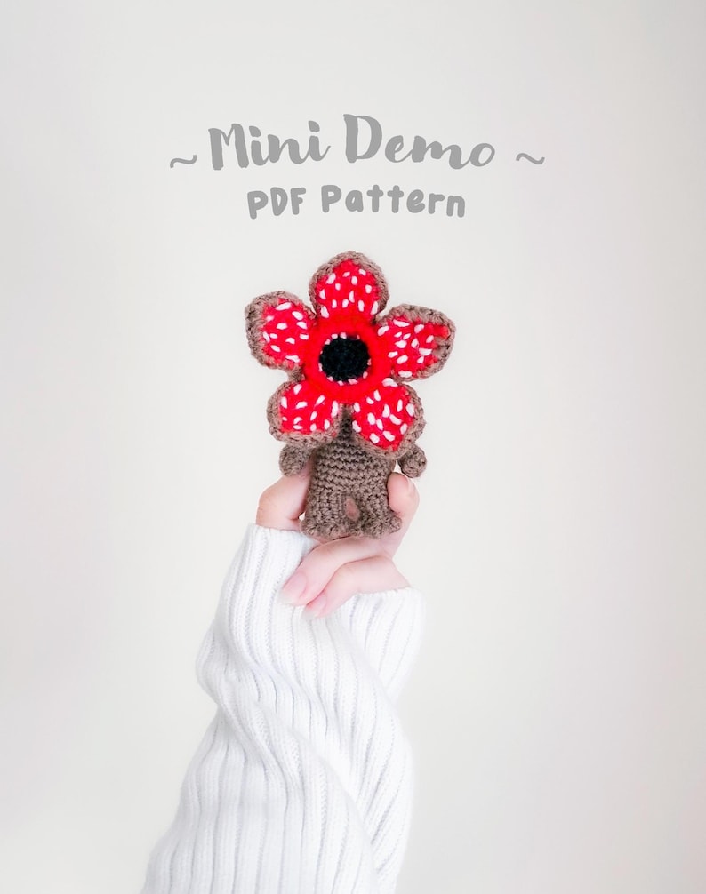 Pode incluir: Um Demogorgon de croch&ecirc;, com uma cabe&ccedil;a vermelha e branca em forma de flor com um centro preto e um corpo marrom. O texto "Mini Demo PDF Pattern" est&aacute; acima do Demogorgon.