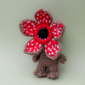 Pode incluir: Um boneco de pel&uacute;cia Demogorgon de croch&ecirc; feito &agrave; m&atilde;o. A criatura tem um corpo, pernas e bra&ccedil;os marrons. A cabe&ccedil;a &eacute; uma grande flor vermelha com manchas brancas e um centro preto. Um item &uacute;nico e artesanal.