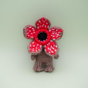 Pode incluir: Figura Demogorgon de croch&ecirc; com uma cabe&ccedil;a vermelha, branca e preta em forma de flor e corpo marrom. O brinquedo de pel&uacute;cia feito &agrave; m&atilde;o tem um design detalhado, com uma apar&ecirc;ncia texturizada e uma est&eacute;tica caprichosa. A figura tem aproximadamente 13 cm de altura.