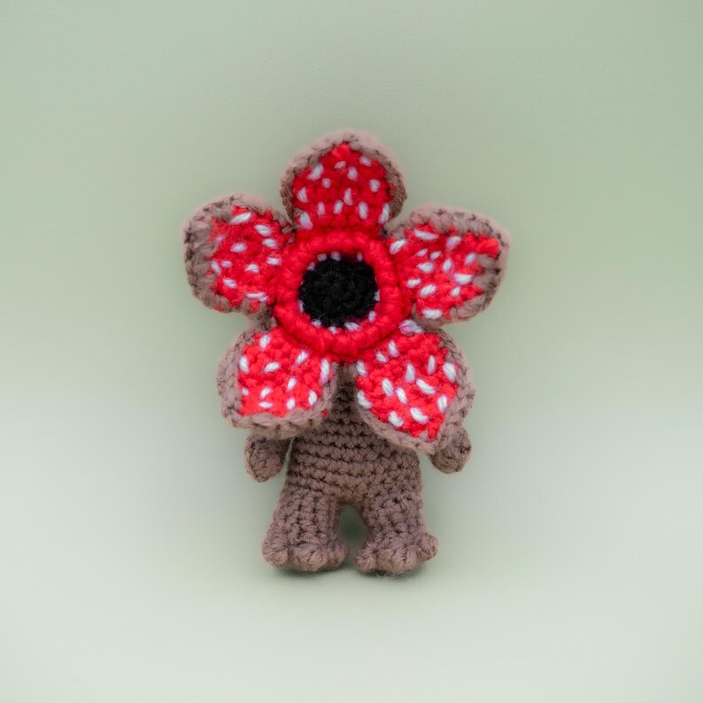 Pode incluir: Figura Demogorgon de croch&ecirc; com uma cabe&ccedil;a vermelha, branca e preta em forma de flor e corpo marrom. O brinquedo de pel&uacute;cia feito &agrave; m&atilde;o tem um design detalhado, com uma apar&ecirc;ncia texturizada e uma est&eacute;tica caprichosa. A figura tem aproximadamente 13 cm de altura.