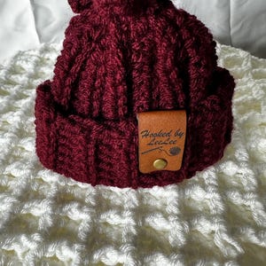 May include: A burgundy crocheted beanie with a pom pom on top and a brown leather tag that says "Hooked by LeeLee".