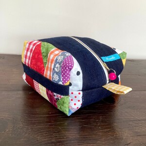 May include: A patchwork denim zippered pouch with a bright pink zipper pull and a yellow and white gingham fabric handle. The pouch is made of denim and has a patchwork design with various colors and patterns, including red, green, yellow, blue, and white.