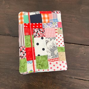 Könnte beinhalten: Eine Patchwork-Zeitschriftenhülle mit einer Vielzahl von Stoffmustern, darunter Tupfen, Blumenmuster und Vichy-Karo. Der Umschlag wird von einem roten Gummiband zusammengehalten. Die Stoffe sind in Rot-, Grün-, Weiß- und Blautönen gehalten.