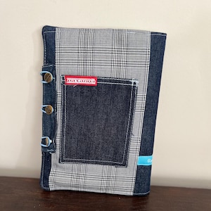 Könnte beinhalten: Ein grauer und weißer karierter Notizbuchumschlag mit einer blauen Jeans-Tasche und einem roten Etikett mit der Aufschrift "paxanga". Der Umschlag hat blaue Knöpfe an der Seite.