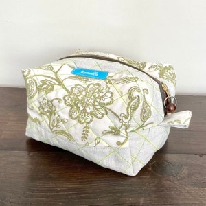 Puede incluir: Una bolsa de maquillaje acolchada blanca con un estampado floral verde y una etiqueta azul que dice "Romanillo".