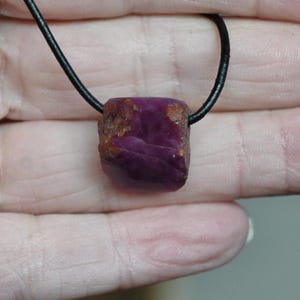 Op de afbeelding: Een ruwe, diep magenta robijn hanger aan een zwart koord. De robijn heeft een ruwe, rechthoekige vorm met natuurlijke texturen en een paar bruine vlekken. Het koord lijkt van leer of een vergelijkbaar materiaal te zijn.