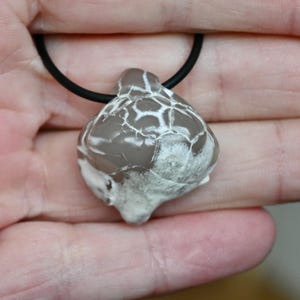 Könnte beinhalten: Halskette mit einem tropfenförmigen Anhänger aus grauem und weißem Stein. Der Stein hat ein rissiges Muster und ist an einer schwarzen Kordel befestigt. Der Anhänger wird in einer Hand gehalten.