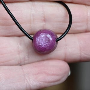 Può includere: Primo piano di una collana con un pendente di pietra preziosa rosa scuro, lucido e rotondo. Il pendente è infilato su un cordino nero. La pietra preziosa ha una superficie strutturata ed è il punto focale della collana.