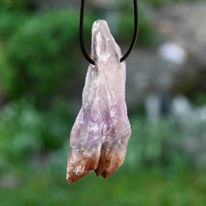 May include: A purple and brown amethyst crystal pendant on a black cord necklace. The crystal is raw and has a natural, pointed shape.