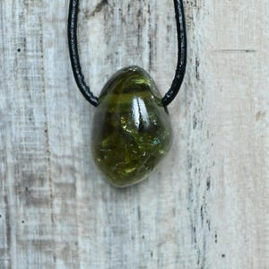 May include: A green gemstone pendant suspended on a black cord. The polished, oval-shaped stone has a translucent quality, with visible internal patterns and a glossy surface. The cord is knotted at the top, ready to be worn.