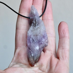 May include: A purple amethyst crystal pendant on a black cord necklace. The crystal is a raw, natural shape with a pointed tip and a flat base.